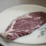 Close-up of a deer backstrap submerged in a glass bowl of milk, with soft lighting highlighting the textures.